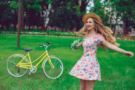 Beautiful blond woman in a straw hat on a lawn with flowers whirls and dances in the park. Bicycle sport and active rest.の写真素材
