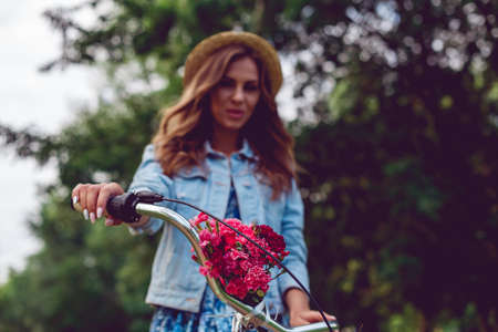 Close-up of a bicycle rudder and blurred background with a young woman in a straw hat in the park. Bouquet of red flowers on the bike.の写真素材