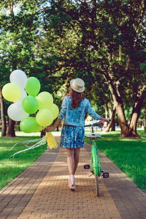 View from the back of a beautiful woman goes with a retro bicycle. Girl in dress and straw hat. Active lifestyle in the park.の写真素材