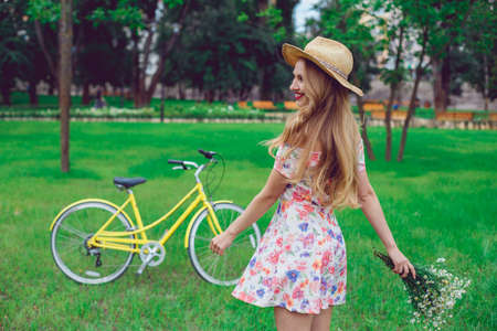 Beautiful happy young woman enjoying summer with wild-flower bunch. Happy girl dancing outdoor, relaxation and good mood in park, sunset. Bicycle on the background.の写真素材