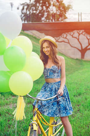 Portrait of a beautiful young caucasian woman on her bike smiling and laughing outdoor. Enjoy summer day, posing on the green lawn. woman in a straw hat holding balloonsの写真素材