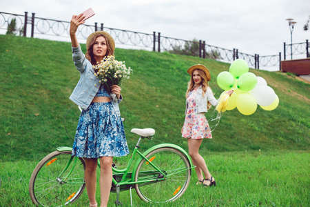 Two young beautiful women posing and making selfies in the park. Girls have fun with a bicycle and balloons.の写真素材