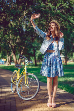 Beautiful young woman in blue dress and a straw hat is doing selfie using a smart phone and smiling while standing near her bicycle in the park.の写真素材