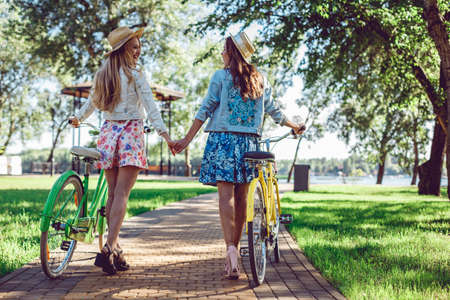 Rear view of two women holding hands walking in a park with bicycles. Best friends spend time actively, sports.の写真素材