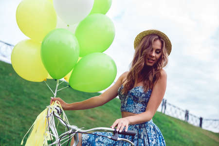 Portait of beautiful woman wearing a nice blue dress having fun in park with bicycle and ballons. Vintage scenery. Pretty girl with retro look, bike and ballons.の写真素材