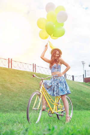 Happy girl posing on yellow bicycle with ballons and smiling. Full length portrait of a smiling woman standing near bicycle on the green lawn.の写真素材