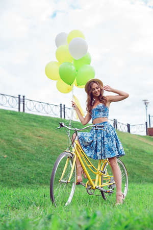Beautiful young woman riding bicycle with hat on her head enjoying summer time. The girl holds the balloons in the color of her bicycle.の写真素材