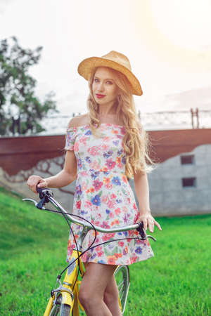 Portrait of beautiful blonde girl riding bike in park. Wearing stylish dress and straw hat. Enjoy summer dayの写真素材