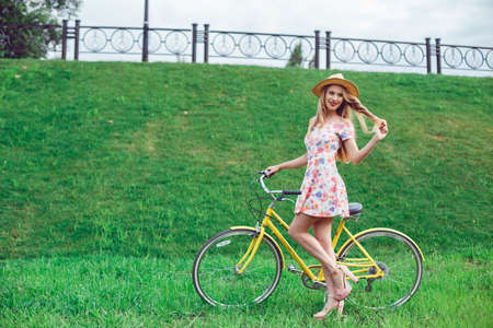 Full length portrait of a beautiful blond woman posing near a yellow bicycle on a green lawn park background. Wearing stylish dress and straw hat. Enjoy summer day,の写真素材