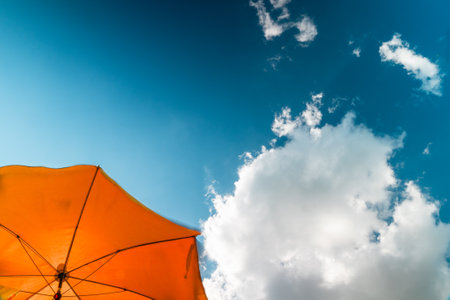 Blue Sky With Beautiful White Clouds Topping an Orange Sun Umbrella Indicating a Sunny Vacation Day on the Beach on a Hot Summer Dayの写真素材