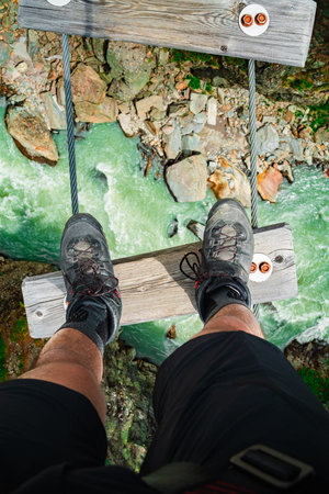 Legs of a Hiker Standing on a Wooden Plank of a Hanging Bridge Far Above a Raging River on Their Travel Journey Path of an Adventurous Trip Top Down Viewの写真素材