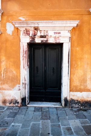 A beautiful black door set in a weathered Venetian building with warm orange walls, capturing the charm and history of architecture. Stone pavement enhances the rustic atmosphere.の写真素材