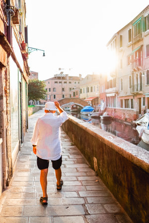 An elderly man strolls beside a charming canal in Venice, Italy, at sunset. He walks leisurely in casual attire, surrounded by water and historic architecture bathed in warm sunlight.の写真素材
