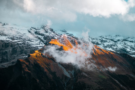Stunning mountain landscape with snow-capped peaks, glowing autumn colors, and dynamic clouds creating a dramatic atmosphere. Inspiration for nature enthusiasts loving breathtaking scenery.の写真素材