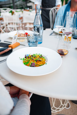 A beautifully presented gourmet risotto dish garnished with microgreens sits on an elegant restaurant table. Nearby, there are glasses of wine, a water bottle, and other dining elements.の写真素材
