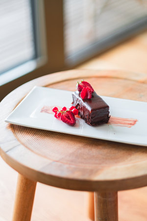 A rich chocolate cake slice garnished with fresh raspberries and currants, elegantly presented on a white plate atop a rustic wooden table, exuding indulgence and gourmet appeal.の写真素材