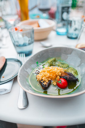A beautifully plated gourmet vegetarian dish featuring a cherry tomato, surrounded by vibrant sauce and garnished with herbs, set in an elegant dining atmosphere with glassware and cutlery.の写真素材