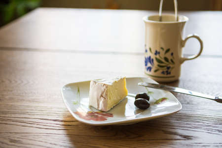 Small depth of field.    A slice of soft cheese and two olives lie on a white plate, on a wooden kitchen tableの写真素材