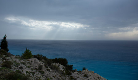 Sun rays from clouds above ocean and island with green trees and bright rocks in Greece.