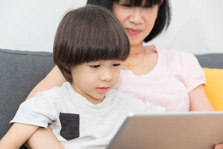 Cheerful Asian mother with little son sitting on sofa and playing video game on tablet while spending time together at home.の写真素材