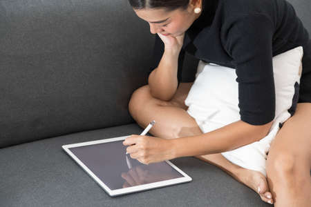 young Asian woman relaxing on sofa writing and looking at digital tablet at home.の写真素材
