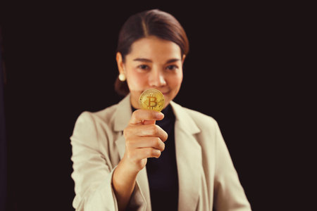 cheerful asian young woman standing on black wall background holding bitcoin looking camera.