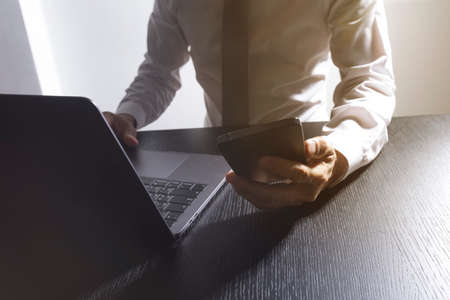 Close-up of the hands of a well-dressed man in a white shirt looking at his smartphone, using a laptop on a desk.の写真素材