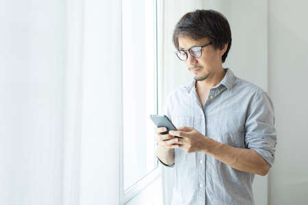 Asian businessman texting on smartphone standing next to window.の写真素材