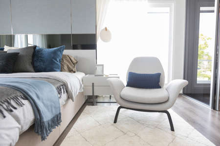 White armchair and carpet next to cozy king size bed with white bedding in fashionable bedroom interior.の写真素材