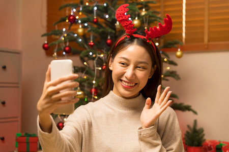 Young Asian woman using smart phone video call talking with friend or boyfriend. Christmas tree decorated with ornament in living room at home. Christmas and New Year holiday festival.の写真素材