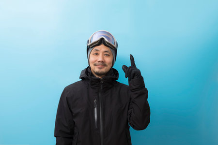 Winter sport. Handsome asian man with snowboard goggles and black anorak coat pointing finger on...