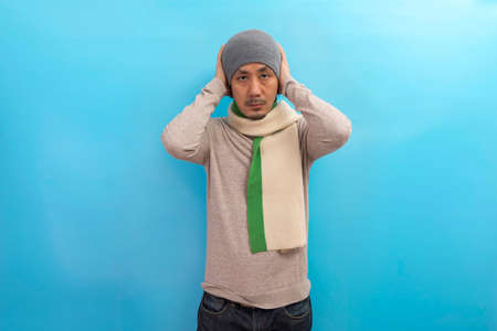 Winter fashion. Handsome asian man in hat and scarf standing and looking straight on blue background.の写真素材