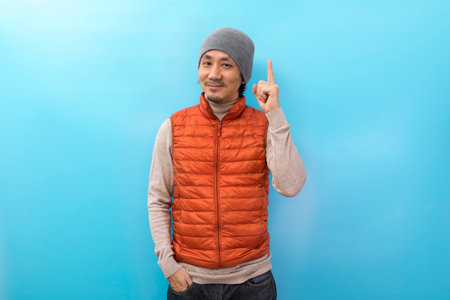 Winter fashion. Handsome asian man with hat and orange jacket pointing finger over white isolated...