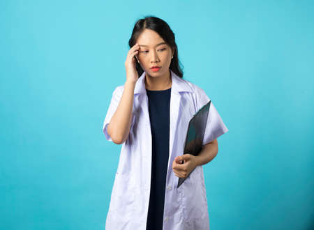 Exhausted young asian female doctor looking tired after shift in hospital, looking sad with fatigue, standing on blue background.の写真素材