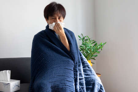 Asian man sick or ill and coughing by covering their mouth with napkin paper while sitting on the sofa at home.の写真素材