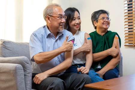 Senior Asian couple and young woman get vaccinated with bandage on arm show thumb up sign in living room. Grandfather and grandmother get vaccine.の写真素材