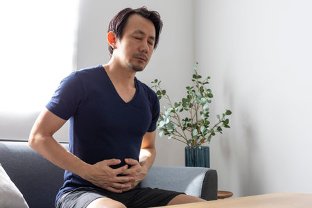 Adult Asian Man Having a Stomach ache Sitting On Sofa At Home.