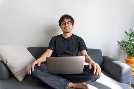 Adult handsome man making video call with friends via laptop while sitting on sofa at his modern home.の写真素材