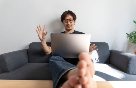 Adult handsome man making video call with friends via laptop while sitting on sofa at his modern home.の写真素材