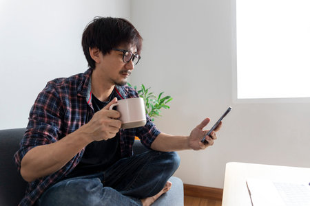 Happy Asian man using smartphone or tablet smiling and relax on sofa at home, man using cellphone...