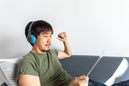 Asian man sitting on sofa while listening to music from wireless headphone or watch online movie...
