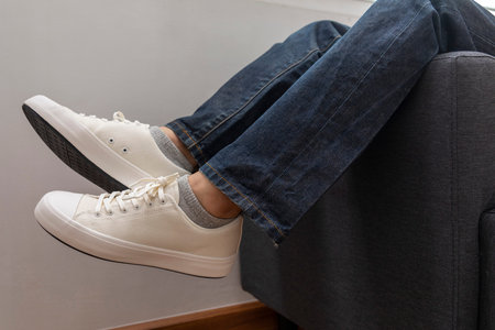 close up of blue jean and white shoe, man relaxing on sofa in a room.