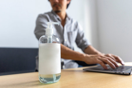 Hand sanitizer gel standing on wooden table. Adult Asian man cleaning hand with sanitizer gel while using laptop computer.の写真素材
