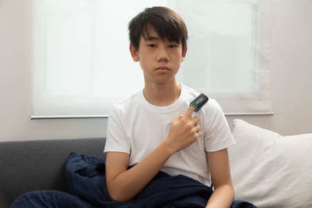Sick asian teen boy with pulse oximeter on his finger. Ill child lying on sofa with blanket in living room.の写真素材