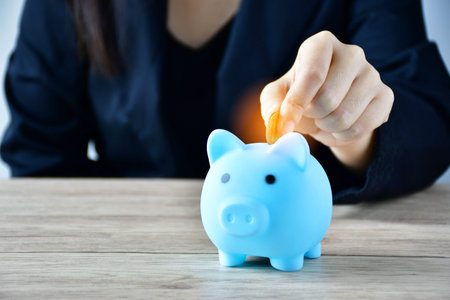 A woman puts a coin in a blue piggy bank on a wooden table to save money And record household income...
