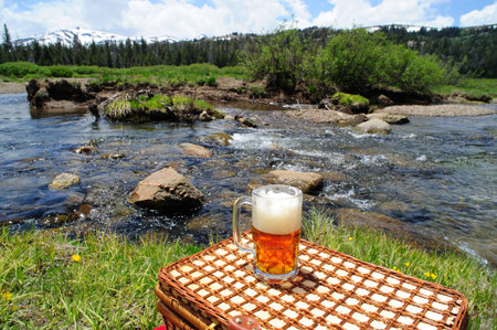 Partially consumed mug of ice cold  micro brewery Ale sitting on a picnic basket used for a table wth crystal clear river and forest in the background.の写真素材