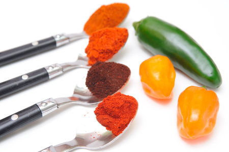 Four different types of chile powder lined up in spoons with fresh jalapeno and habanero peppers....