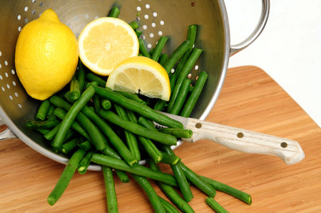 Green beans with a sliced lemon in a stainless steel colander with a small knife and bamboo cutting...
