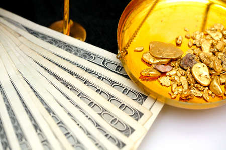 Gold nuggets in the pan of a balance scale with many fifty and hundred dollar bills showing the value of bothの写真素材
