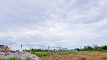 View of an open field surrounded by a metal security fence under a cloudy sky. The ground has patches of dry grass and mounds of sand or light-colored soil.の写真素材